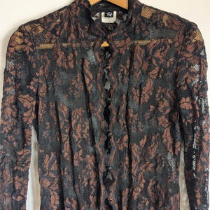 May include: A long-sleeved shirt made of black and copper-coloured floral lace. The garment features a high collar, a front button-down closure with small round buttons, and a label at the neckline with the letters TD printed in white.