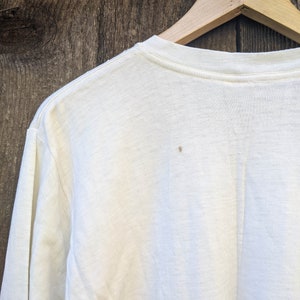 May include: A white long-sleeved t-shirt with a crew neck. The shirt is hanging on a wooden hanger against a woodgrain background.
