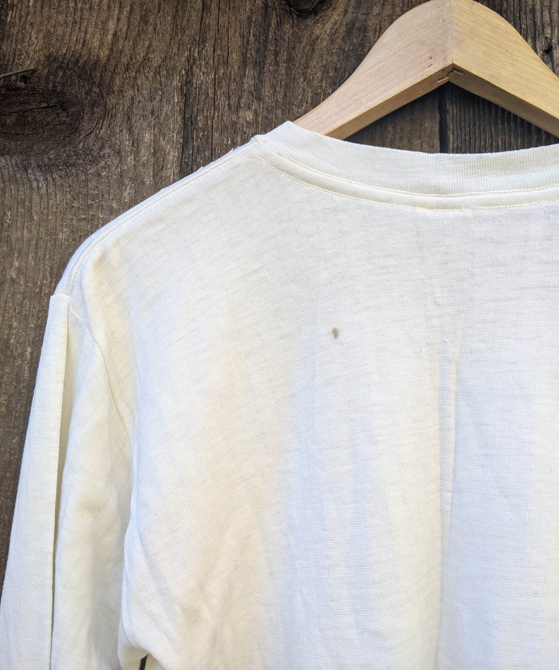 May include: A white long-sleeved t-shirt with a crew neck. The shirt is hanging on a wooden hanger against a woodgrain background.