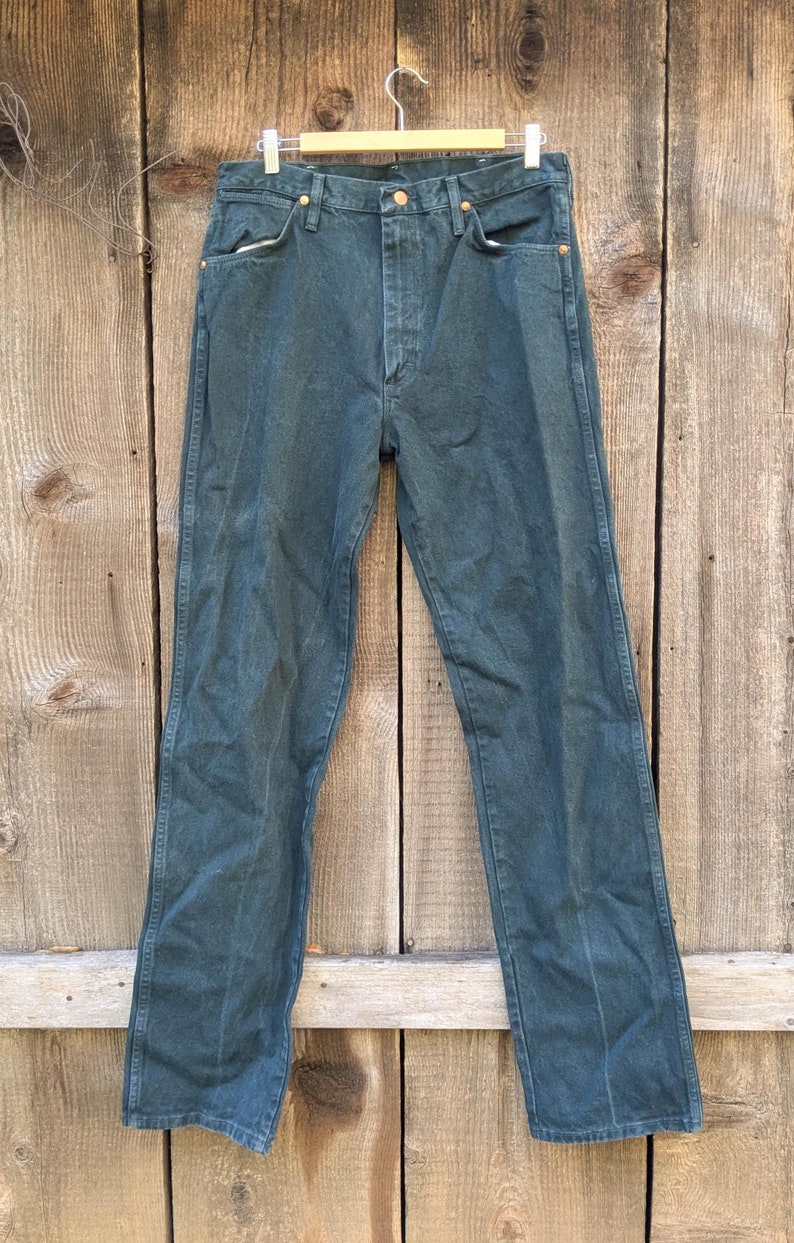 May include: A pair of dark green denim jeans hanging on a wooden hanger against a wood panel background.