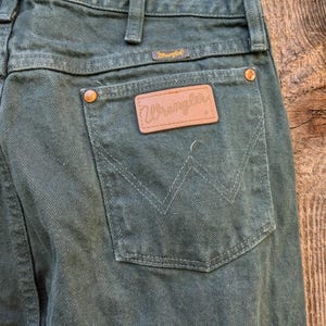 May include: A pair of dark green Wrangler jeans with a brown leather patch on the back pocket that says "Wrangler".