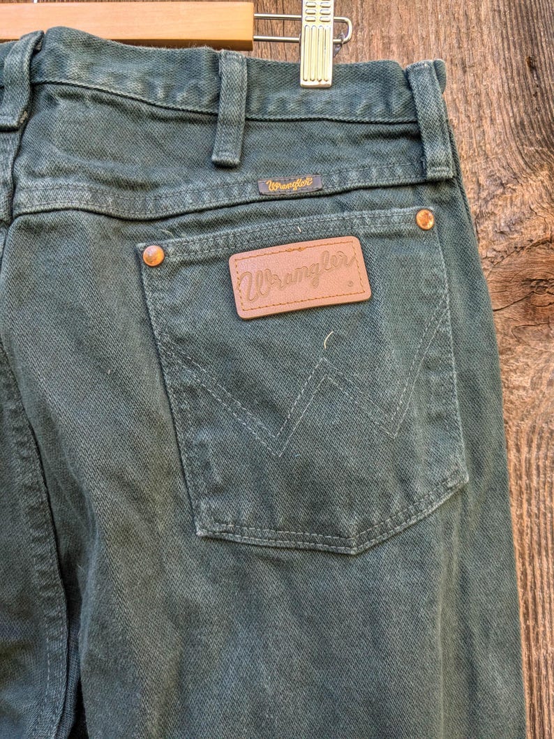 May include: A pair of dark green Wrangler jeans with a brown leather patch on the back pocket that says "Wrangler".