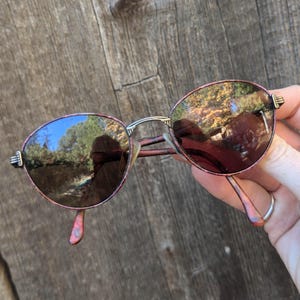 May include: A pair of oval-shaped sunglasses with a silver frame and brown lenses. The frame has a pink marbled pattern.
