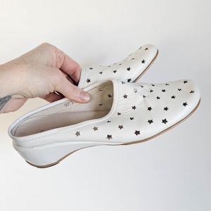 May include: White leather clogs with star-shaped cutouts. The shoes have a low wedge heel and a slip-on design. The clogs are held by a hand against a white background. The shoes are approximately 9 inches long.