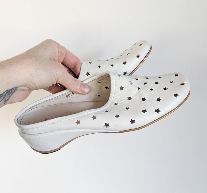 May include: White leather clogs with star-shaped cutouts. The shoes have a low wedge heel and a slip-on design. The clogs are held by a hand against a white background. The shoes are approximately 9 inches long.