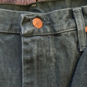 May include: Close-up of a dark green pair of jeans with a copper button. The button has the word "Wrangler" embossed on it.