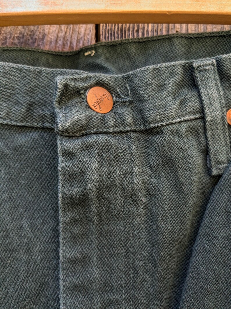 May include: Close-up of a dark green pair of jeans with a copper button. The button has the word "Wrangler" embossed on it.