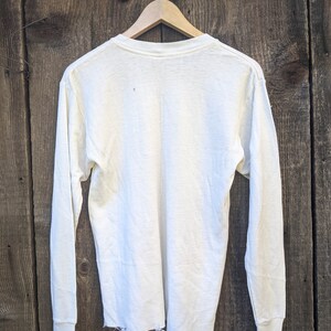May include: A white long-sleeved t-shirt with a distressed hem. The shirt is hanging on a wooden hanger against a wood background.