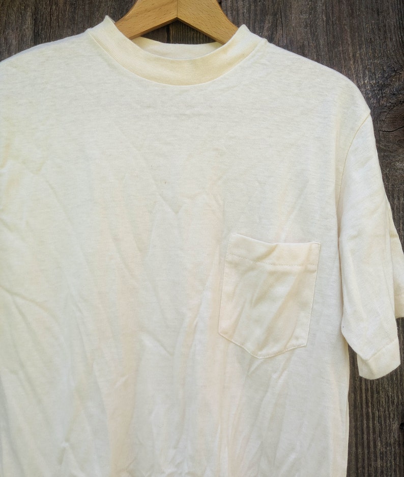 May include: A plain white t-shirt with a single pocket on the left side. The shirt is made of a soft, comfortable fabric and has a crew neck.