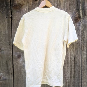 May include: A plain, off-white t-shirt hanging on a wooden hanger against a woodgrain background.