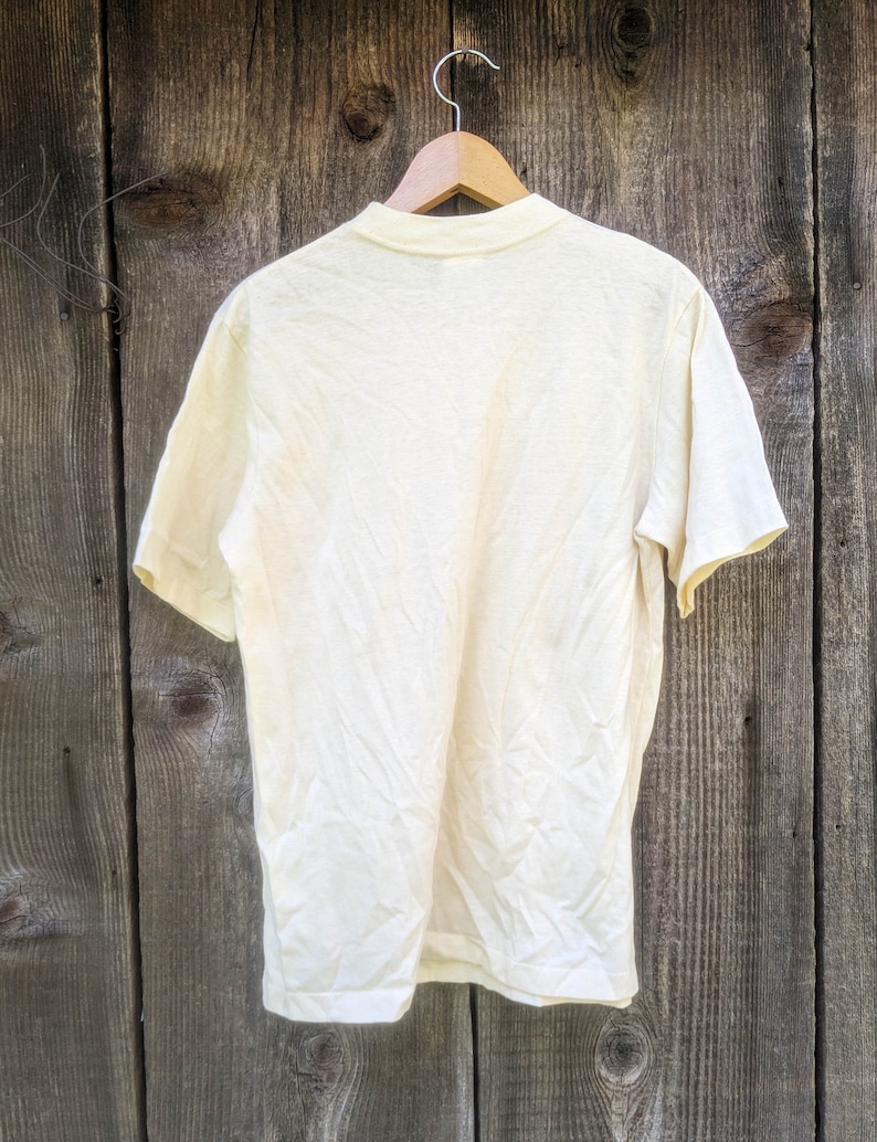 May include: A plain, off-white t-shirt hanging on a wooden hanger against a woodgrain background.