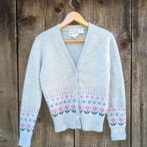 May include: A light blue cardigan sweater with a V-neck and button closure. The sweater features a floral pattern in pink, purple, and green at the bottom. The sweater is hanging on a wooden hanger.