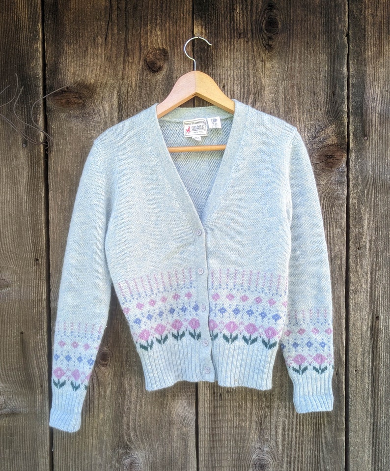 May include: A light blue cardigan sweater with a V-neck and button closure. The sweater features a floral pattern in pink, purple, and green at the bottom. The sweater is hanging on a wooden hanger.