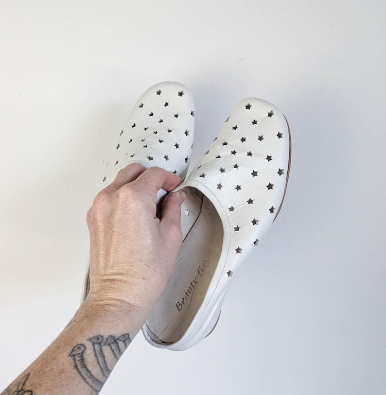 May include: A pair of white leather slip-on shoes with star-shaped cutouts. The shoes have a rounded toe and a low heel. The brand name "Beauty fis" is visible on the inner sole.