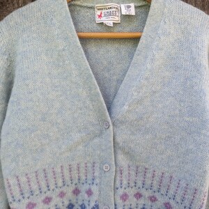 May include: A light blue cardigan sweater with a V-neck and button closure. The sweater features a floral pattern in shades of purple and blue at the bottom. The label reads "Shetlander Crazy Horse 100% Wool".