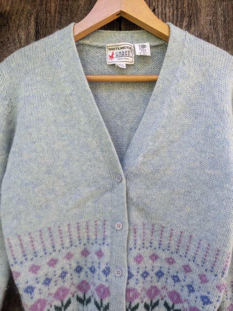 May include: A light blue cardigan sweater with a V-neck and button closure. The sweater features a floral pattern in shades of purple and blue at the bottom. The label reads "Shetlander Crazy Horse 100% Wool".