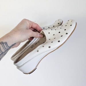 May include: Cream-colored slip-on shoes with star cutouts. The shoes have a wedge heel and a rounded toe. The sole is brown, and the heel is approximately 2 inches high. The shoes are held by a hand.