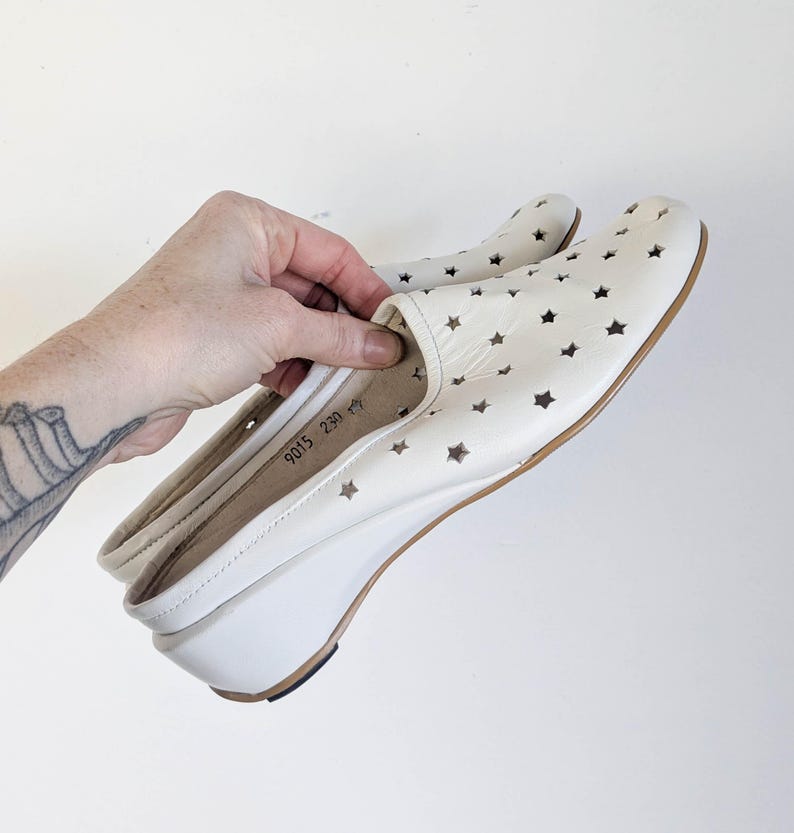 May include: Cream-colored slip-on shoes with star cutouts. The shoes have a wedge heel and a rounded toe. The sole is brown, and the heel is approximately 2 inches high. The shoes are held by a hand.