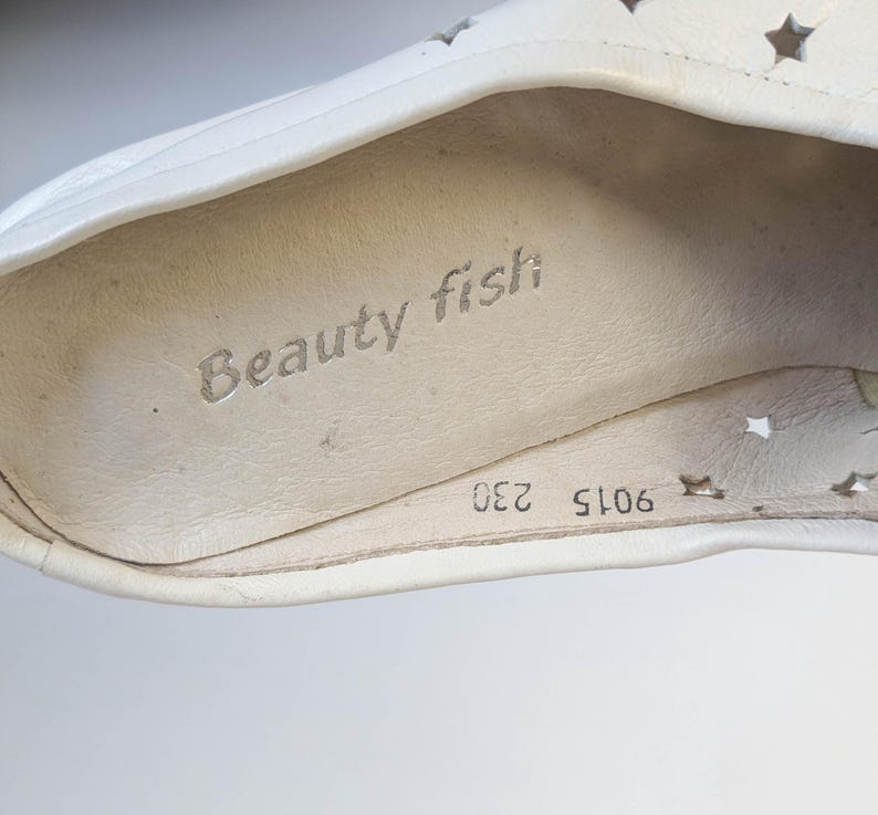 May include: Close-up of a white shoe interior with the text "Beauty fish" printed in gray. The shoe has star-shaped cutouts and the numbers "9015 230" are visible.