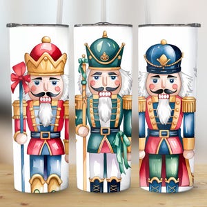 May include: Three white tumblers feature watercolor illustrations of nutcrackers in red, green, and blue uniforms. Each nutcracker has a white beard, a mustache, and a decorative hat. The tumblers are cylindrical and appear to be made of stainless steel.