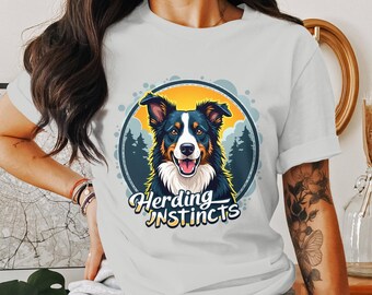 以下が含まれることがあります： 黒と白のボーダーコリー犬のグラフィックが描かれた白いTシャツ。犬は、黄色い太陽と緑の木々のある青い円の中にいます。犬の下には「Herding Instincts」という文字が印刷されています。