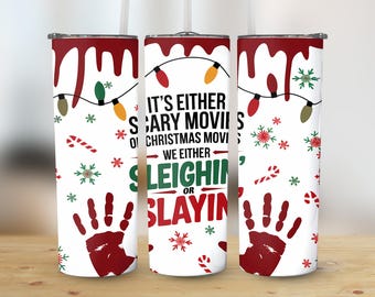 May include: A white tumbler with a red lid and straw features a festive design. The design includes a dripping red border, Christmas lights, snowflakes, candy canes, and handprints. Text reads "It's Either Scary Movies or Christmas Movies, We Either Sleighin' or Slayin'."