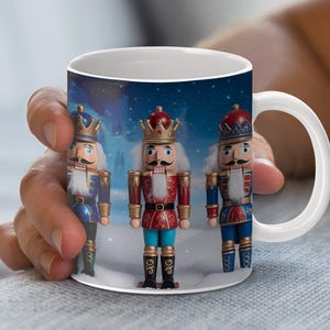 May include: White ceramic mug featuring three toy nutcrackers in festive attire. The nutcrackers are in red and blue uniforms with gold accents, set against a snowy winter scene. The mug is held by a person.