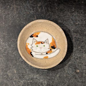 May include: A small ceramic bowl featuring a calico cat design. The cat is curled up in a ball with its eyes closed. The bowl is beige with brown speckles.