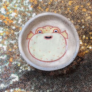 May include: A small, round ceramic dish featuring a hand-painted pufferfish design. The pufferfish has a cream-coloured body, red outline, and yellow accents. The dish is beige with a speckled texture, ideal for holding small items.