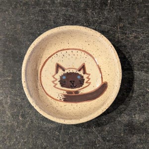 May include: A small ceramic bowl with a speckled beige glaze. The bowl features a hand-painted design of a brown and white cat with blue eyes.