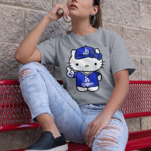 May include: A grey t-shirt featuring a cartoon Hello Kitty wearing a blue Dodgers baseball cap and jersey. The shirt has the text "LA Dodgers" on the jersey. The person is wearing ripped blue jeans and black shoes.
