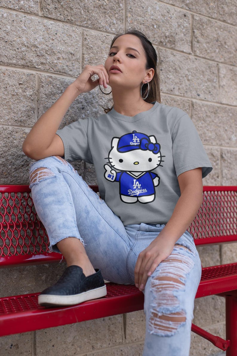 May include: A grey t-shirt featuring a cartoon Hello Kitty wearing a blue Dodgers baseball cap and jersey. The shirt has the text "LA Dodgers" on the jersey. The person is wearing ripped blue jeans and black shoes.