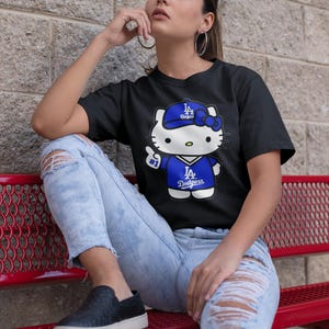 May include: Black t-shirt featuring a cartoon Hello Kitty character wearing a Los Angeles Dodgers baseball cap and jersey. The character is giving a thumbs up. The shirt is paired with ripped blue jeans.