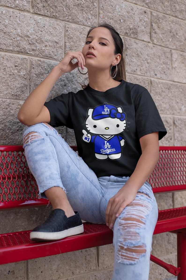May include: Black t-shirt featuring a cartoon Hello Kitty character wearing a Los Angeles Dodgers baseball cap and jersey. The character is giving a thumbs up. The shirt is paired with ripped blue jeans.