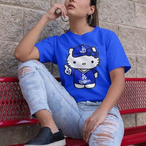 May include: Royal blue t-shirt featuring a cartoon Hello Kitty character wearing a Los Angeles Dodgers baseball cap and jersey. The graphic includes the Dodgers logo and the number 1. The shirt is paired with distressed blue jeans.