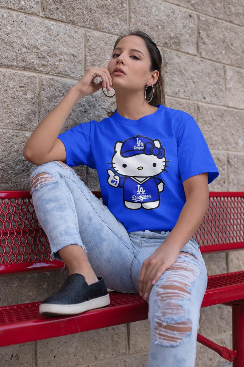 May include: Royal blue t-shirt featuring a cartoon Hello Kitty character wearing a Los Angeles Dodgers baseball cap and jersey. The graphic includes the Dodgers logo and the number 1. The shirt is paired with distressed blue jeans.