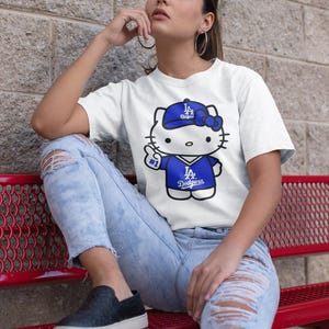 May include: White t-shirt featuring a cartoon Hello Kitty character wearing a blue Dodgers baseball cap and jersey. The character is giving a thumbs up. The shirt has the text "#1" and "Dodgers".