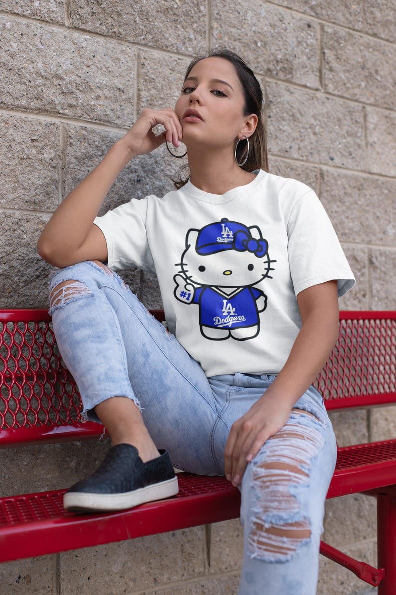 May include: White t-shirt featuring a cartoon Hello Kitty character wearing a blue Dodgers baseball cap and jersey. The character is giving a thumbs up. The shirt has the text "#1" and "Dodgers".