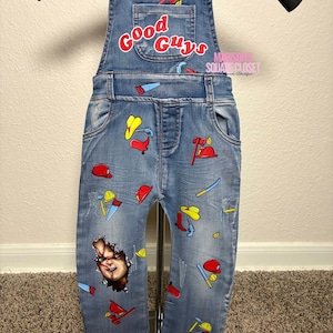 May include: Light blue denim overalls with the text "Good Guys" in red and white on the bib pocket. The overalls are decorated with colorful cartoon images of tools and a Chucky graphic. The overalls are hanging on a black hanger.