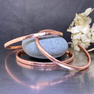 Peut inclure: Trois bracelets en cuivre martelé empilés sur une pierre grise. Les bracelets sont de tailles différentes et ont une finition brillante.