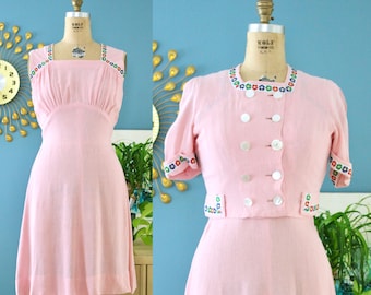 May include: A pink dress with a white trim and colourful floral embroidery. The dress has a square neckline and a fitted bodice. The skirt is full and flows out from the waist. The dress is made of a lightweight fabric and is perfect for summer.
