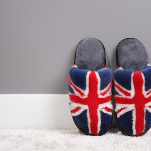 May include: A pair of plush slippers featuring the Union Jack flag design. The slippers have a navy blue base with a red and white flag pattern. The interior and top are a soft gray material.