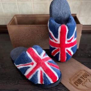 May include: A pair of blue, red, and white Union Jack flag slippers. The slippers have a dark gray interior and a black sole. The slippers are made of a soft, furry material. The slippers are displayed on a dark brown wooden surface.