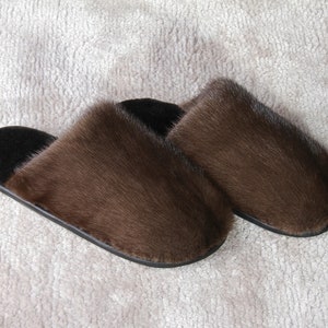May include: A pair of brown fur slippers with black soles. The slippers have a plush, textured appearance and are designed for indoor wear. The fur is a rich, warm brown color, and the soles are a contrasting black.