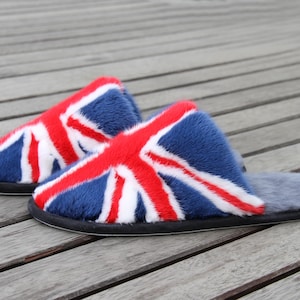 May include: A pair of plush slippers featuring the Union Jack flag design. The slippers have a blue base with red and white accents, a black sole, and a gray interior. The slippers are made of a soft, furry material.