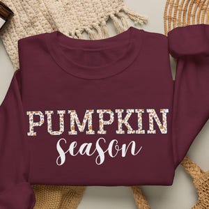 May include: A maroon long-sleeve crewneck sweatshirt featuring the text PUMPKIN SEASON in white. The word PUMPKIN is filled with a small floral and pumpkin pattern, while the word season is written in a white cursive script font.