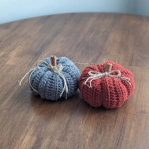 May include: Two crocheted pumpkins in shades of blue and red-orange sit on a wooden surface. Each pumpkin has a cinnamon stick stem and is tied with twine. The pumpkins are handmade and decorative.