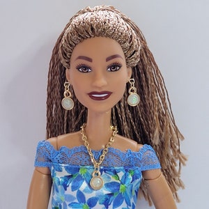 May include: A doll with brown skin and long brown braids wearing a blue and white floral dress with a lace trim. She is wearing gold earrings and a gold necklace with a white pendant.
