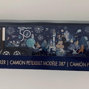 May include: A gold Peterbilt Model 387 hauler toy truck in its original packaging. The trailer features a dark blue design with Walt Disney World 50th anniversary graphics, including fireworks, a castle, and various Disney characters.