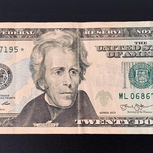 Pode incluir: Uma nota de vinte dólares com um retrato de Andrew Jackson. A nota apresenta um selo verde e números de série, com o texto "The United States of America" e "Twenty Dollars". A nota retangular é de cor creme.